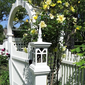 May include: White picket fence with a white wooden archway covered in yellow roses. A white wooden post with a decorative lantern top is attached to the fence.