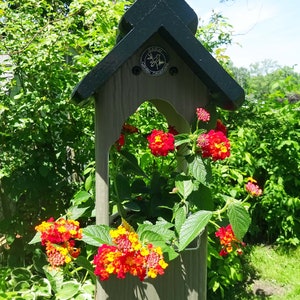 May include: A hanging planter with a gray wooden frame and a black roof. The planter is filled with a vibrant orange and red flowering plant. The planter has a metal hook for hanging and a metal tag with the text "Sadler Collection" on it.