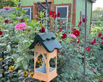 May include: A wooden bird feeder shaped like a house with a green roof hangs from a metal hook. The feeder has a clear plastic insert and is surrounded by a variety of flowers.