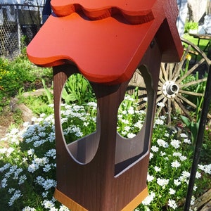 May include: A wooden bird feeder with a red roof and two feeding ports. The feeder is hanging from a chain and is surrounded by white flowers.