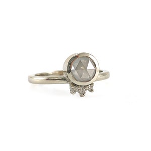 May include: A silver ring with a round, bezel-set, gray diamond. The diamond has a faceted cut. The ring has a band of silver and a row of smaller diamonds set in a row below the main diamond.