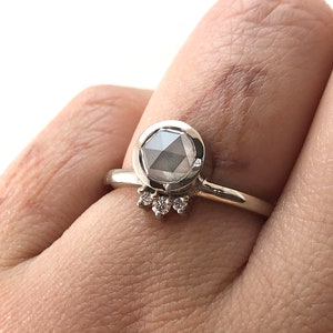 May include: A silver ring with a large, round, gray gemstone set in a bezel setting. The ring has a delicate band and a row of smaller diamonds set in a halo around the gemstone.