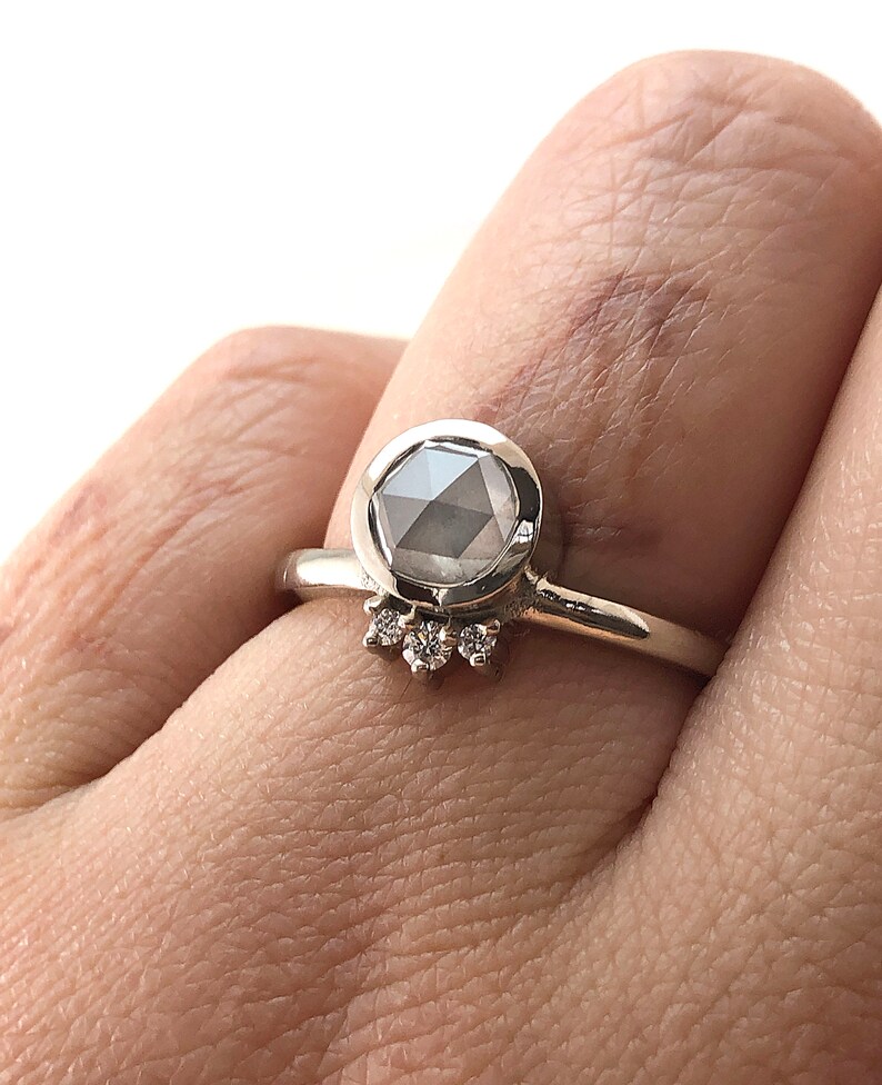 May include: A silver ring with a large, round, gray gemstone set in a bezel setting. The ring has a delicate band and a row of smaller diamonds set in a halo around the gemstone.