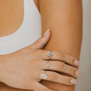 May include: A hand wearing four gold rings. The rings feature a variety of designs, including a round stone, a small cluster of stones, a simple band, and a religious medallion.