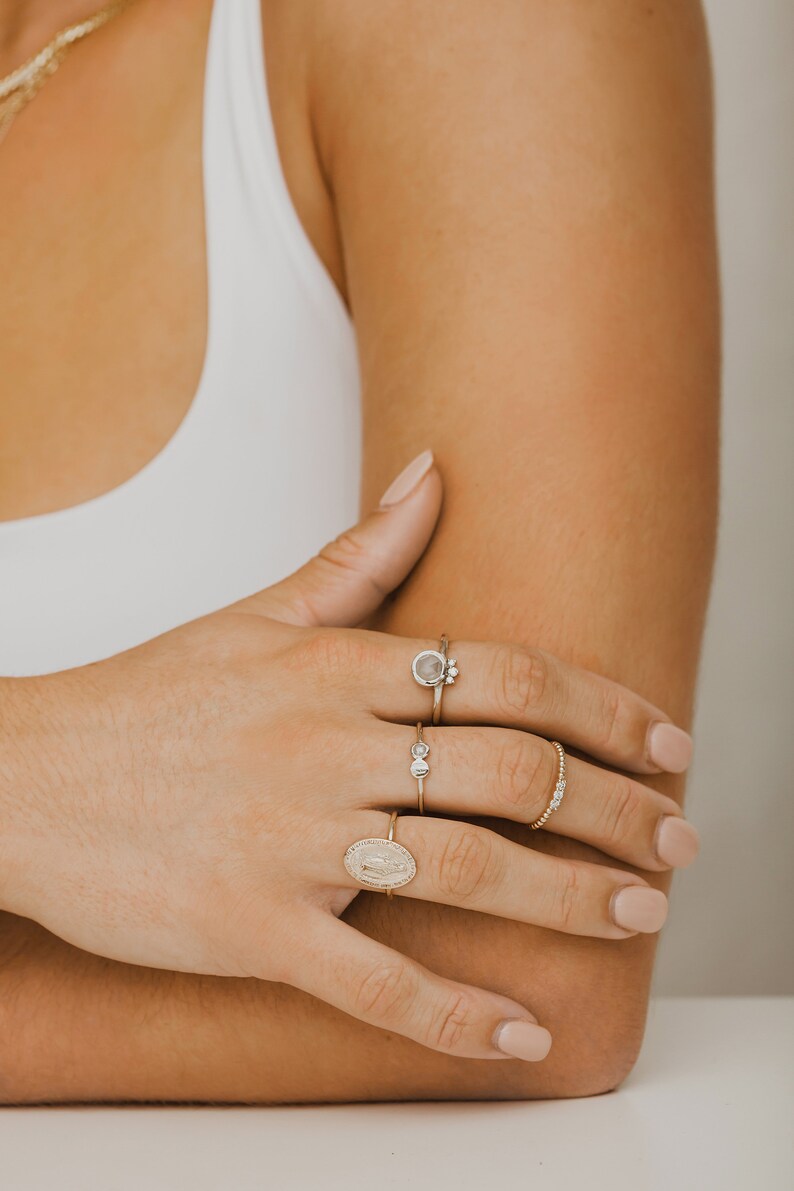 May include: A hand wearing four gold rings. The rings feature a variety of designs, including a round stone, a small cluster of stones, a simple band, and a religious medallion.