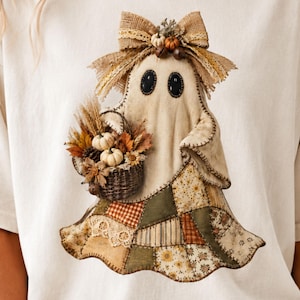 May include: A cream-colored t-shirt featuring a graphic of a ghost wearing a burlap bow and holding a basket filled with miniature pumpkins, wheat, and autumn leaves. The ghost has a patchwork-style skirt made of various brown, green, and tan fabrics.