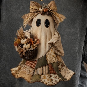 May include: A charcoal grey t-shirt featuring a graphic of a ghost wearing a large tan bow and holding a woven basket filled with miniature pumpkins, dried wheat, and autumn leaves. The ghost has a patchwork skirt in shades of olive, tan, and brown.