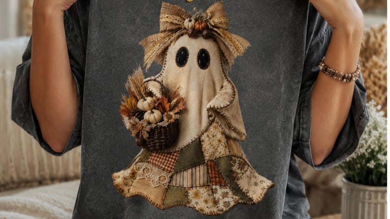 May include: A charcoal grey t-shirt featuring a graphic of a ghost wearing a large tan bow and holding a woven basket filled with miniature pumpkins, dried wheat, and autumn leaves. The ghost has a patchwork skirt in shades of olive, tan, and brown.
