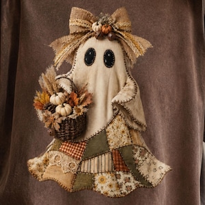 May include: A dark brown t-shirt featuring a central graphic of a ghost figure. The ghost has black oval eyes, a hessian bow on its head, and carries a small woven basket filled with miniature white pumpkins, dried wheat, and autumn leaves.