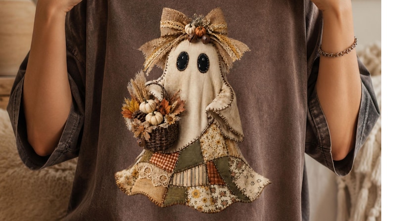 May include: A dark brown t-shirt featuring a central graphic of a ghost figure. The ghost has black oval eyes, a hessian bow on its head, and carries a small woven basket filled with miniature white pumpkins, dried wheat, and autumn leaves.