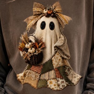 May include: A charcoal grey sweatshirt featuring a central graphic of a ghost figure. The ghost has black oval eyes, a hessian bow on its head, and holds a dark wicker basket filled with miniature pumpkins, wheat, and autumn leaves.