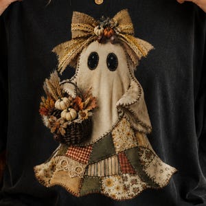 May include: A black t-shirt featuring a graphic of a ghost wearing a hessian bow and holding a basket filled with small pumpkins, wheat, and autumn leaves. The ghost has a patchwork-style lower body with various brown, green, and cream patterns.