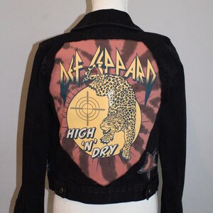 May include: A black denim jacket featuring a large back patch with a tie-dye background. The graphic displays a leopard, a target symbol, and the text Def Leppard High 'N' Dry. A small star-shaped patch is visible on the lower right side of the jacket.