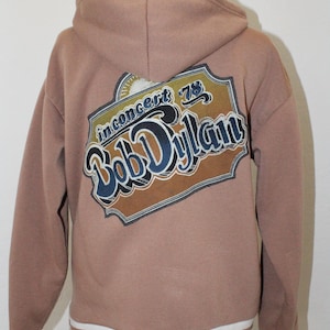 May include: A tan hooded sweatshirt featuring a graphic on the back with the text "Bob Dylan" and "in concert 78" inside a decorative border. The garment has long sleeves with white and tan striped ribbed cuffs and a matching striped hem.