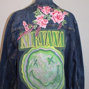 May include: A dark blue denim jacket featuring a large embroidered patch on the back. The patch displays the word NIRVANA in yellow letters above a green, distorted smiley face with X-shaped eyes. Pink floral embroidery sits above the patch.