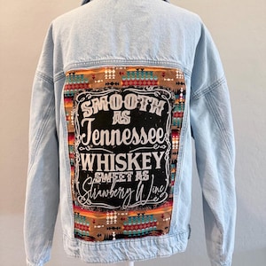 May include: A light blue denim jacket featuring a large rectangular back panel with a brown background and a colorful geometric pattern. The center of the panel displays the text: Smooth as Tennessee Whiskey Sweet as Strawberry Wine.