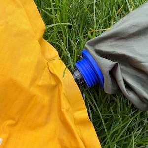 Op de afbeelding: Close-up van een gele en olijfgroene kampeerwaterzak op groen gras. De zak heeft een blauwe schroefdop. Het gele gedeelte is een heldere, effen kleur. Het olijfgroene gedeelte is een donkerdere, gedempte kleur.