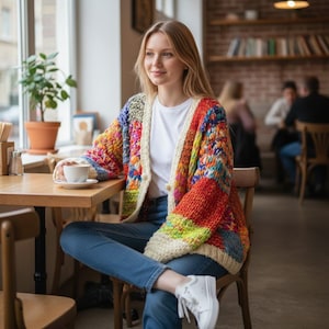 May include: A colorful, chunky knit cardigan with a patchwork design. The sweater features a mix of vibrant colors including red, orange, yellow, green, blue, and purple. It has a cream-colored border and buttons. The person is wearing a white t-shirt and blue jeans.