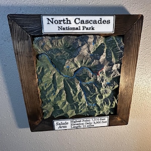 May include: A 3D topographic map of North Cascades National Park encased in a dark stained wooden frame. The map features green terrain with blue water elements. Labels read North Cascades National Park and Sahale Arm Highest Point: 7,570 feet.