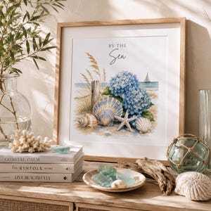 Könnte beinhalten: Ein quadratischer Kunstdruck in einem Rahmen mit einer Aquarellillustration von blauen Hortensien, Muscheln, einem Seestern und Strandgras an einem sandigen Ufer mit einem Segelboot am Horizont. Der Text BY THE Sea ist oben mittig in Schwarz gedruckt.
