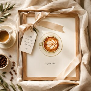 K&ouml;nnte beinhalten: Ein gerahmter Aquarelldruck eines Latte in einer Tasse mit Untertasse und dem Text 'espresso' darunter. Ein beiges Band ist um den Rahmen gebunden, an dem ein Schild mit der Aufschrift 'a little something for your new beginning' und einem kleinen Herzsymbol h&auml;ngt.