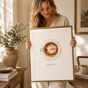 K&ouml;nnte beinhalten: Ein gerahmter Kunstdruck mit einer Aquarellillustration einer Kaffeetasse mit Latte-Art-Motiv in der Mitte. Die Tasse ist aus der Vogelperspektive dargestellt und das Wort espresso ist in einer einfachen schwarzen Schrift unter der Tasse gedruckt.