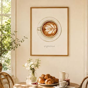 K&ouml;nnte beinhalten: Ein gerahmter Wandkunstdruck mit einer Aquarellillustration einer Kaffeetasse mit Latte-Art von oben, zentriert &uuml;ber dem Wort espresso. Das Kunstwerk ist in einem hellen Holzrahmen an einer Wand &uuml;ber einem Tisch mit Speisen und Blumen zu sehen.