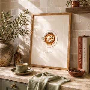 K&ouml;nnte beinhalten: Ein gerahmter Kunstdruck mit einer Aquarellillustration einer Espressotasse mit Latte-Art-Muster, zentriert &uuml;ber dem Wort espresso. Der Druck befindet sich in einem hellen Holzrahmen mit wei&szlig;em Passepartout und steht auf einer Arbeitsplatte.