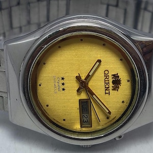 May include: An Orient wristwatch with a round gold-toned dial, silver-toned hands, and a day-date window at the three o'clock position. The dial features the text Orient, Crystal, and 21 Jewels with three stars. It has a silver-toned metal link band.