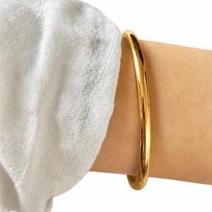 May include: A polished gold-tone bangle bracelet with a smooth, rounded surface. The bangle features a continuous, solid circular design with a high-shine reflective finish that catches the light, creating a clean and simple aesthetic for the wrist.