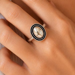 May include: A silver-toned ring featuring an oval-shaped centre piece. The centre is a smooth, polished silver oval surrounded by a textured black border with small silver beads along the outer edge, set on a simple, polished silver band.
