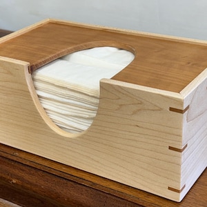 May include: A wooden tissue box with a light-colored body and a darker, contrasting top. The box has a curved opening for tissue access and is filled with white tissues. The corners are reinforced with small, decorative wooden accents.