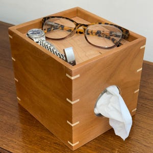 May include: A wooden tissue box with a watch, eyeglasses, and a silver object inside. The box is a light brown color with white accents at the corners. White tissues are visible coming out of the circular opening.