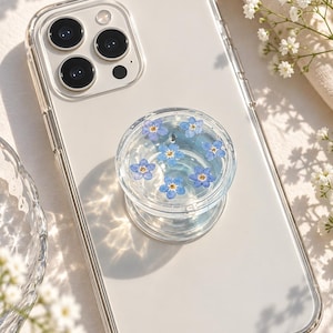 May include: A clear smartphone case with a circular collapsible phone grip attached to the back. The grip features a transparent resin top embedded with several small, light blue forget-me-not flowers with yellow centres, creating a floral design.