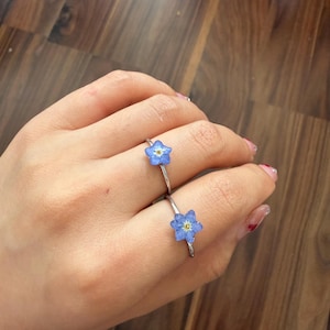 May include: Two silver-toned rings, each featuring a small, five-petaled blue flower with a yellow center. The flowers appear to be preserved or pressed, and each is securely mounted onto a thin, polished metal band worn on the fingers.