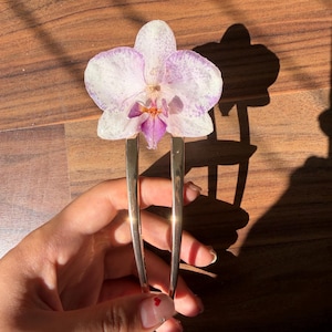 May include: A gold-toned metal hair fork featuring a pressed orchid flower at the top. The orchid has white petals with light purple speckles and a deep purple and orange center. The hair fork has two smooth, polished prongs extending downward.