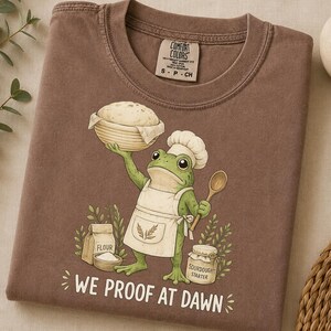 Könnte beinhalten: Ein braunes T-Shirt mit einer Grafik eines grünen Frosches, der eine Kochmütze und eine Schürze trägt und eine Schüssel Teig sowie einen Holzlöffel hält. Darunter sind ein Mehlbeutel und ein Glas Sourdough Starter sowie der Text WE PROOF AT DAWN.