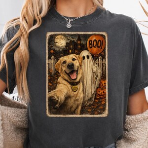 May include: A charcoal gray t-shirt featuring a graphic print of a golden retriever wearing a tag that says GOOD BOY, standing next to a ghost holding an orange balloon with the text BOO, with a haunted house and pumpkins in the background.
