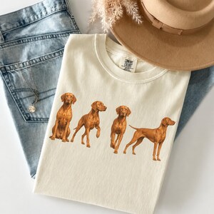Può includere: Una t-shirt color crema con una stampa grafica di quattro cani Vizsla in varie pose. La maglia presenta un girocollo e un'etichetta del marchio Comfort Colors all'interno del colletto. Il design raffigura i cani in calde tonalità marrone dorato.