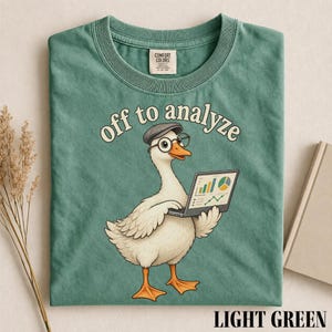 Könnte beinhalten: Ein hellgrünes T-Shirt mit einer Grafik einer weißen Ente, die eine graue Mütze und eine runde Brille trägt und einen Laptop mit bunten Geschäftsgrafiken hält. Der Text über der Ente lautet "off to analyze" in geschwungener Schrift.