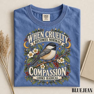 Może przedstawiać: Niebieski t-shirt z grafiką sikorki siedzącej na gałęzi w otoczeniu kwiatów i jagód. Tekst głosi: When cruelty becomes normal, compassion looks radical. Koszulka posiada metkę marki Comfort Colors przy kołnierzyku.