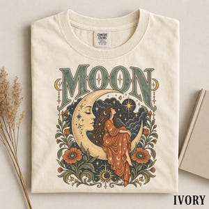 Könnte beinhalten: Ein elfenbeinfarbenes T-Shirt mit einem Grafikdruck einer Person mit langem Haar, die auf einer Mondsichel sitzt. Das Wort MOON ist in großen, stilisierten Buchstaben darüber gedruckt. Das Design enthält dunkle Wolken, Sterne und orangefarbene Blumen.
