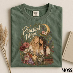 Puede incluir: Una camiseta verde musgo con un gráfico de dos figuras sin rostro rodeadas de flores, una luna creciente, un frasco etiquetado como Rose Petals, una vela, un mortero con mano y libros etiquetados como Herbs, Spells y Remedies con el texto Practical Magic.