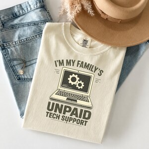 Op de afbeelding: Een crèmekleurig T-shirt met korte mouwen en een afbeelding van een laptop met twee tandwielen op het scherm. De tekst luidt I'M MY FAMILY'S UNPAID TECH SUPPORT. Het shirt ligt op een lichtblauwe spijkerbroek onder een beige vilten hoed.