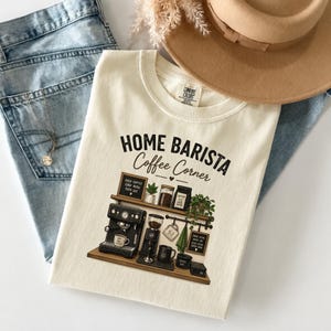 May include: A cream-colored t-shirt featuring a graphic of a coffee station with the text "HOME BARISTA Coffee Corner". The design includes an espresso machine, coffee grinder, mugs, and small signs. The shirt is layered with blue jeans and a tan hat.