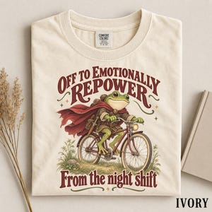 May include: An ivory t-shirt featuring a graphic of a green frog wearing a red cape and brown tunic while riding a bicycle. The text above reads "OFF TO EMOTIONALLY REPOWER" and the text below reads "From the night shift" in a dark red font.