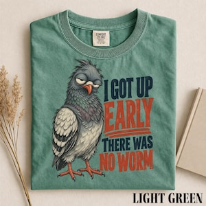 Pode incluir: Uma t-shirt verde-clara com o gráfico de um pombo com aspeto rabugento, penas eriçadas e bico laranja. O texto diz I GOT UP EARLY THERE WAS NO WORM em letras de bloco azul-escuras e vermelhas. A t-shirt tem um decote redondo.