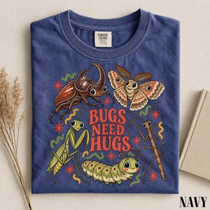 Puede incluir: Una camiseta azul marino de cuello redondo con un estampado gráfico de un escarabajo, una polilla, una mantis religiosa, una oruga y un insecto palo. El texto BUGS NEED HUGS está impreso en naranja. Etiqueta Comfort Colors en el cuello.