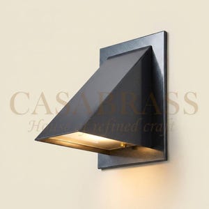 May include: A matte black wall sconce featuring a geometric, triangular housing mounted on a rectangular backplate. The fixture directs light downward from an internal source, creating a warm glow against the wall surface below the unit.