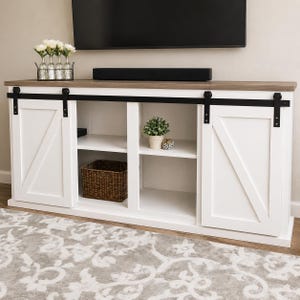 Może przedstawiać: Biało-brązowa szafka RTV z przesuwanymi drzwiami w stylu stodoły. Szafka ma jasnobrązowy blat, czarne okucia i biały korpus. Posiada otwarte półki i pleciony kosz. Widoczna jest mała roślina i elementy dekoracyjne.